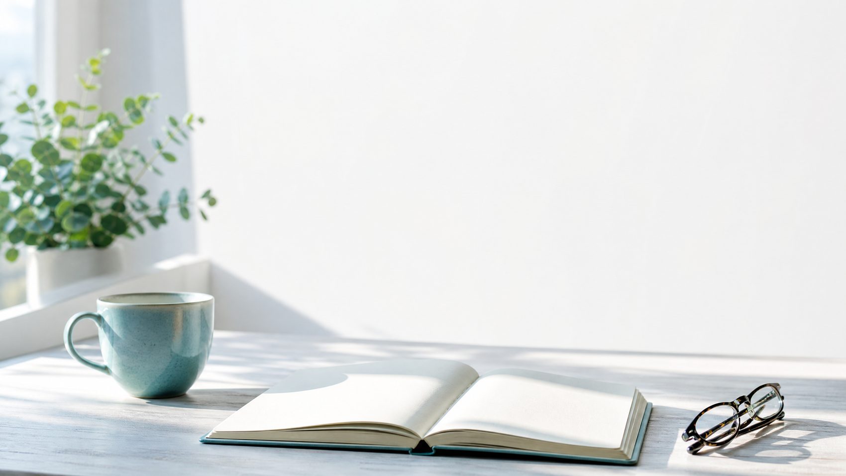 An open blank notebook on a pale desk beside a mug and folded glasses in strong morning light